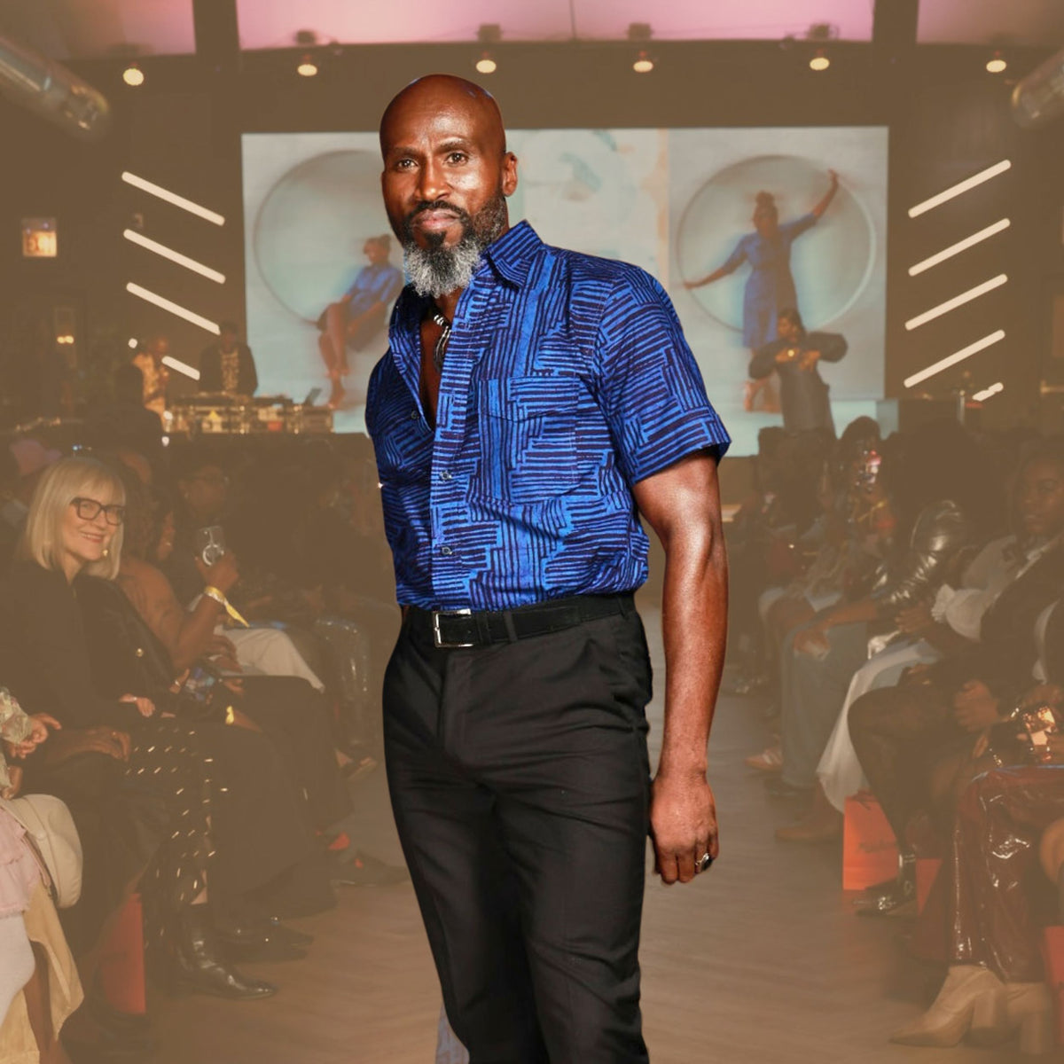 Man walking on a runway at a fashion show with an audience and screen in the background. Each shirt is individually wax-printed, dyed, and stitched by Ghanaian artisans — no two ever the same.