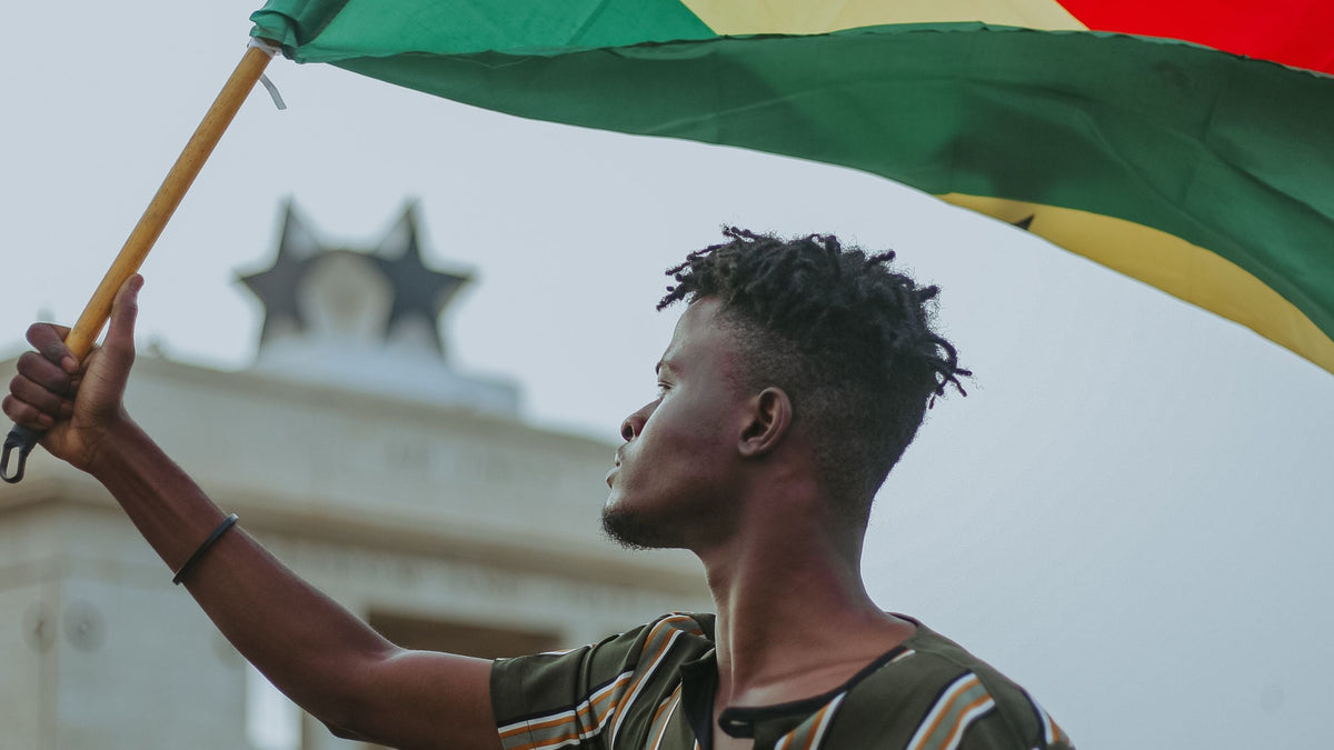 Person holding a flag with a building in the background. Made in Ghana. LOVE DOT INC.