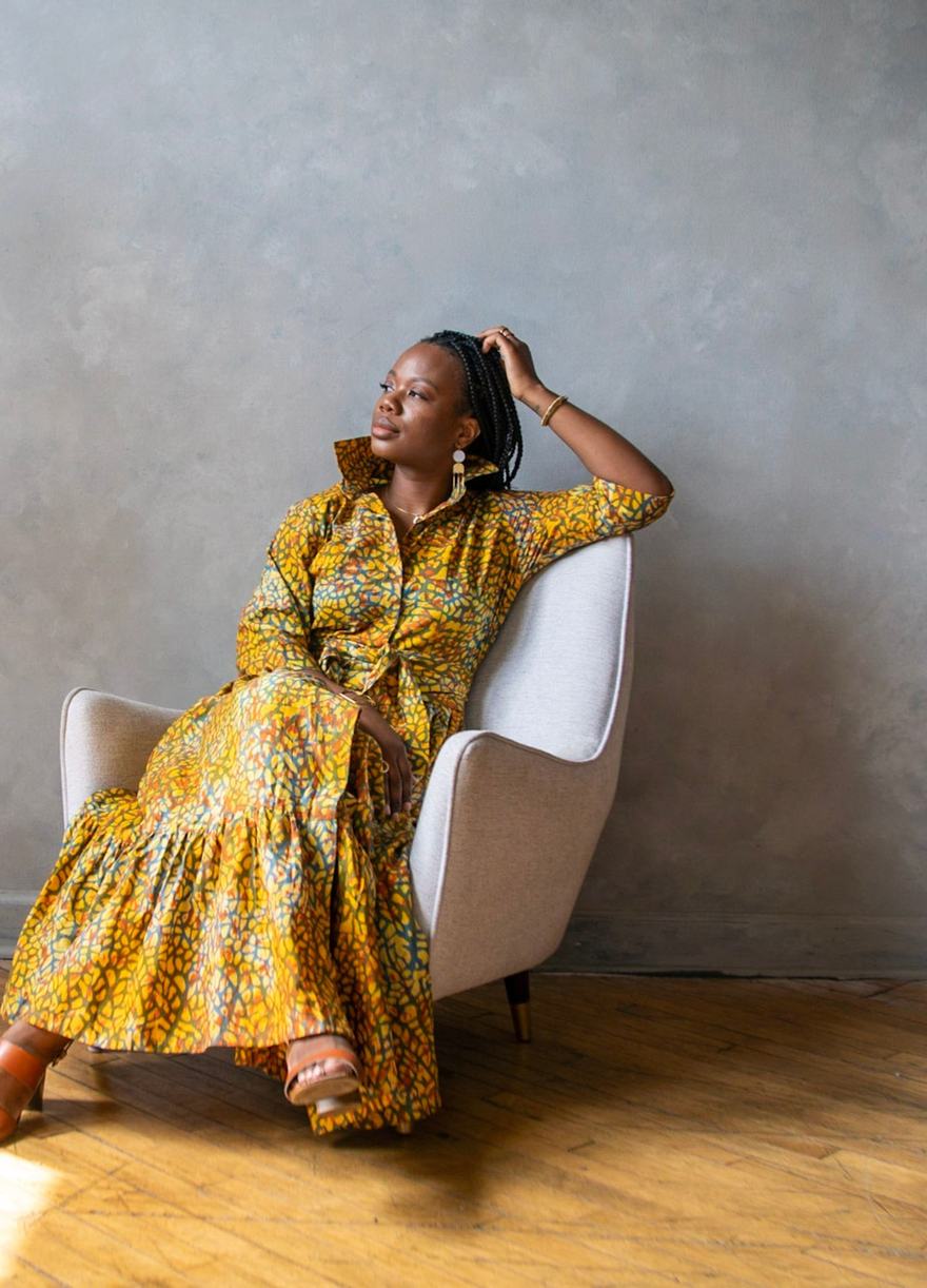 A woman seated in an armchair in a sunlit room, wearing a yellow printed LOVE DOT dress.